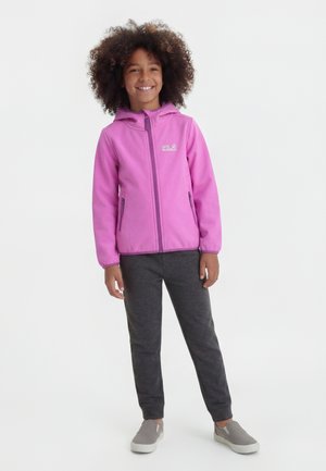 Χαμογελαστό παιδί με σγουρά μαλλιά, που φοράει ροζ ζακέτα με φερμουάρ, σκούρο γκρι παντελόνι και γκρι παπούτσια χωρίς κορδόνια, στέκεται μπροστά σε λευκό φόντο.