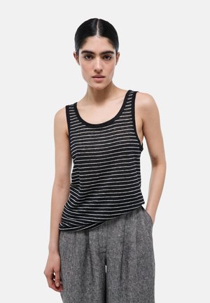 Jeune femme aux cheveux foncés dans un chignon bas portant un haut sans manches rayé noir et blanc et un pantalon gris ample, debout avec une main dans la poche.