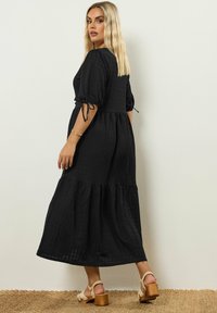Vestido maxi negro en capas con mangas cortas abullonadas, cintura elástica y detalles de lazo. Tela texturizada y dobladillo acampanado. Usado con sandalias de tacón de madera.