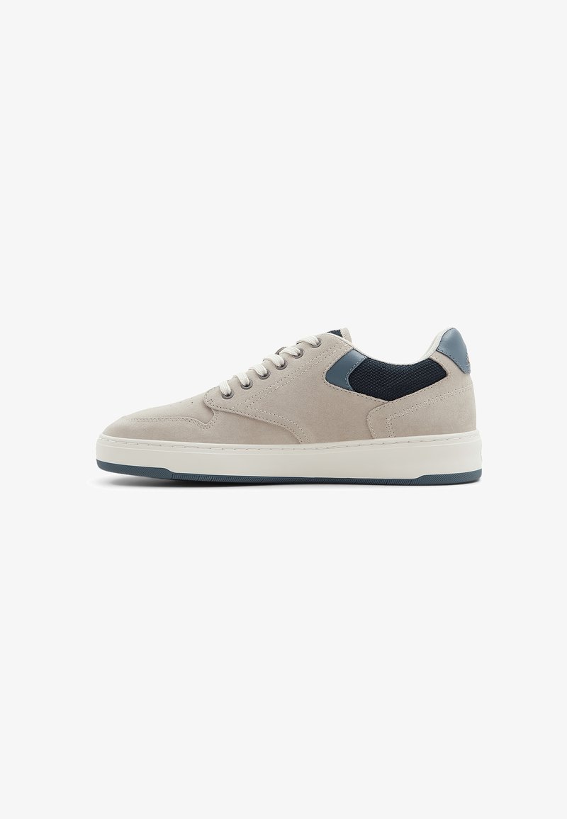 Beige suede sneakers with a textured navy mesh panel, white rubber sole, and grey heel accent. Features lace closure and minimalist design.