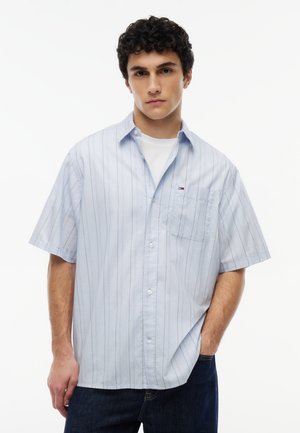 Ung mand med kort krøllet hår iført en lyseblå kortærmet skjorte med striber over en hvid t-shirt, hænderne i lommerne.