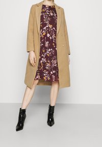 Tammenruskea villakangastakki viininpunaisen kukkakuvioisen mekon päällä, jossa on pinkkejä ja keltaisia yksityiskohtia, yhdistettynä kiiltäviin mustiin nilkkureihin.