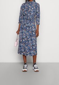 Midi šaty s květinovým vzorem v modré barvě, s páskem v oblasti pasu, límečkem a tříčtvrtečními rukávy, doplněné o černé a bílé tenisky.