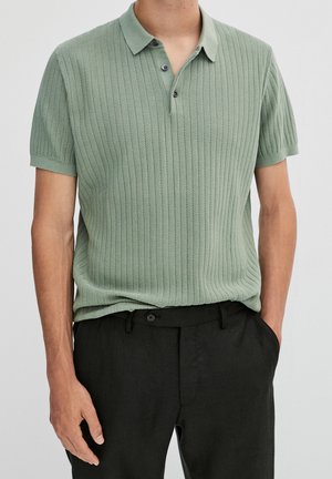 Polo à manches courtes vert clair avec un motif côtelé texturé, un col avec col, et trois boutons foncés, associé à un pantalon noir.