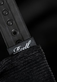 Black corduroy fabric with ridged texture, featuring a black woven label with white "Reell" text and a plastic strap with holes for adjustment.