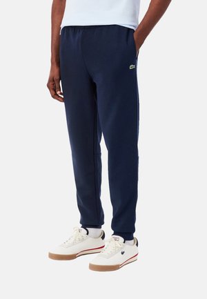 Pantalones deportivos azul marino con cintura elástica, bolsillos laterales y detalle de logo verde. Combinados con zapatillas blancas con suelas color canela y franjas rojas.
