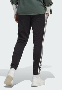 Svarta joggarbyxor med vita sidosträck, elastisk midja, åtsittande mudd, och i kombination med vita sportskor, står på en ljus bakgrund.