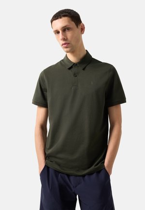 Ung mand iført mørkegrøn kortærmet polo og marineblå bukser, stående med hænderne i lommerne foran en ensartet baggrund.