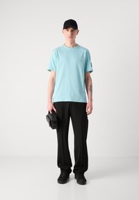 Světle modré tričko s krátkým rukávem, černé kalhoty, černé tenisky a černá čepice. Drží černou tašku s hladkým povrchem. Jednoduchý design.