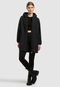 Junge Frau, die eine schwarze Kapuzen-Langjacke, ein schwarzes Crop-Top, schwarze Leggings und schwarze klobige Stiefel trägt und vor einem weißen Hintergrund steht.