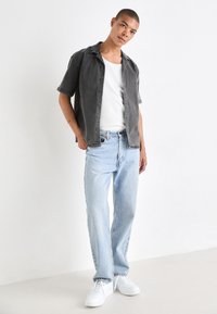 Grå kortermet skjorte over en hvit singlet, kombinert med lyseblå løpetid jeans og hvite joggesko på en ensfarget bakgrunn.
