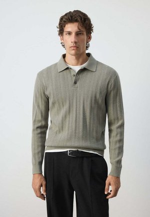 Jeune homme aux cheveux courts et bouclés porte un polo gris texturé à manches longues par-dessus un sous-vêtement blanc et un pantalon noir avec ceinture sur un fond uni.