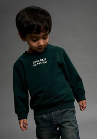Sweat-shirt en coton vert avec des manches longues et des poignets côtelés. Présente un texte blanc indiquant "BONNES JOURS EN ROUTE" centré sur le devant.