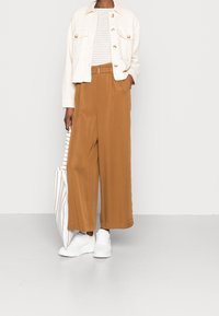 White denim jacket with large buttons, striped shirt, brown wide-leg trousers, and white sneakers. Striped tote bag in hand.