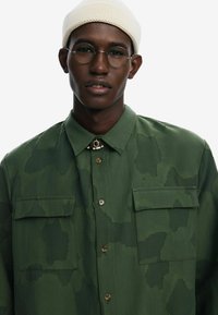 Olive green shirt with a textured camouflage pattern, button front, two chest pockets, and a rounded collar; model wears a white beanie and glasses.