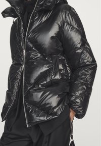 Black puffer jacket with a shiny, quilted design. Features a high collar, zip closure, and a patch pocket on the front. Soft texture.