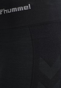 Gros plan sur un tissu noir texturé avec le logo blanc embossé "hummel" et un motif chevrons subtil.