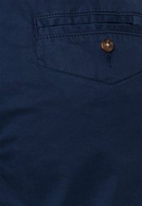 Granatowy materiał o gładkiej fakturze, z tylną kieszenią z brązowym guzikiem oraz kontrastowymi detalami przeszycia.