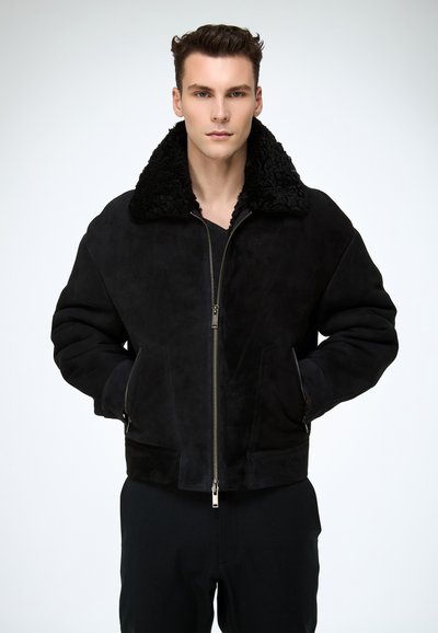 Veste en suède noir avec un col en fausse fourrure noire moelleuse, fermeture éclair et poches latérales. Caractérisée par une texture douce et un design structuré.