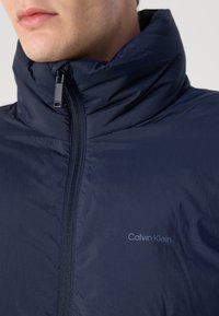 Marineblauwe pufferjack met hoge kraag, ritsluiting en een subtiele "Calvin Klein" logo op de linkerborst, met een soepele, lichte textuur.