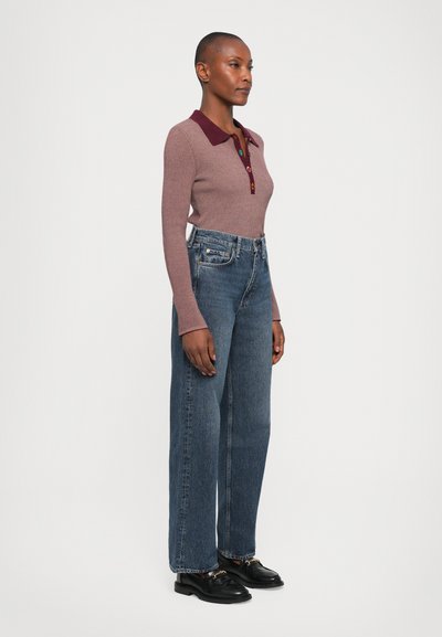 Kvinne som står i en ribbet langermet polo topp med flerfargede knapper, høymidje vide jeans og svarte loafers med gulldetaljer.