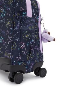 Trolley-Koffer aus dunkelblauem Stoff mit bunten Blumenmustern, lila Akzenten und einem flauschigen Schlüsselanhänger. Enthält zwei schwarze Räder und Griffe.