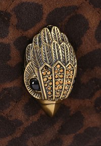 Ciondolo decorativo a forma di testa di gufo in oro e nero con piume testurizzate e rhinestone ambra su uno sfondo di tessuto a pois marrone e nero.
