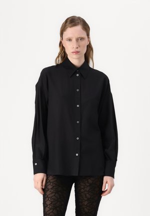 Svart skjorte med knapper og lange ermer, med klassisk krage og subtil glans. Festet med runde svarte knapper. Teksturerte svarte leggings.