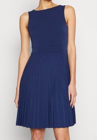 Robe sans manches bleu marine avec un corsage ajusté et une jupe évasée plissée. Fabriquée en tissu lisse, avec un col arrondi et une taille sans coutures.