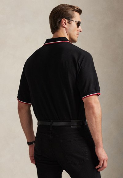 Férfi fekete galléros ingben, piros és fehér szegéllyel, fekete nadrágban, napszemüvegben és órával, háttal egy egyszínű háttér előtt.