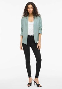 Femme avec de longs cheveux bouclés, portant un blazer vert clair, un haut blanc, un jean skinny noir et des mules à talons hauts noires, se tenant contre un fond blanc.