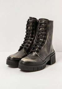 Une paire de bottines noires en cuir à lacets, avec des semelles à crampons épaisses et des talons carrés, sur un fond uni.