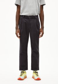 Corduroy broek in donkergrijs met een rechte pijp, plooien aan de voorkant en een knoopsluiting, gecombineerd met levendige gele en oranje sneakers.