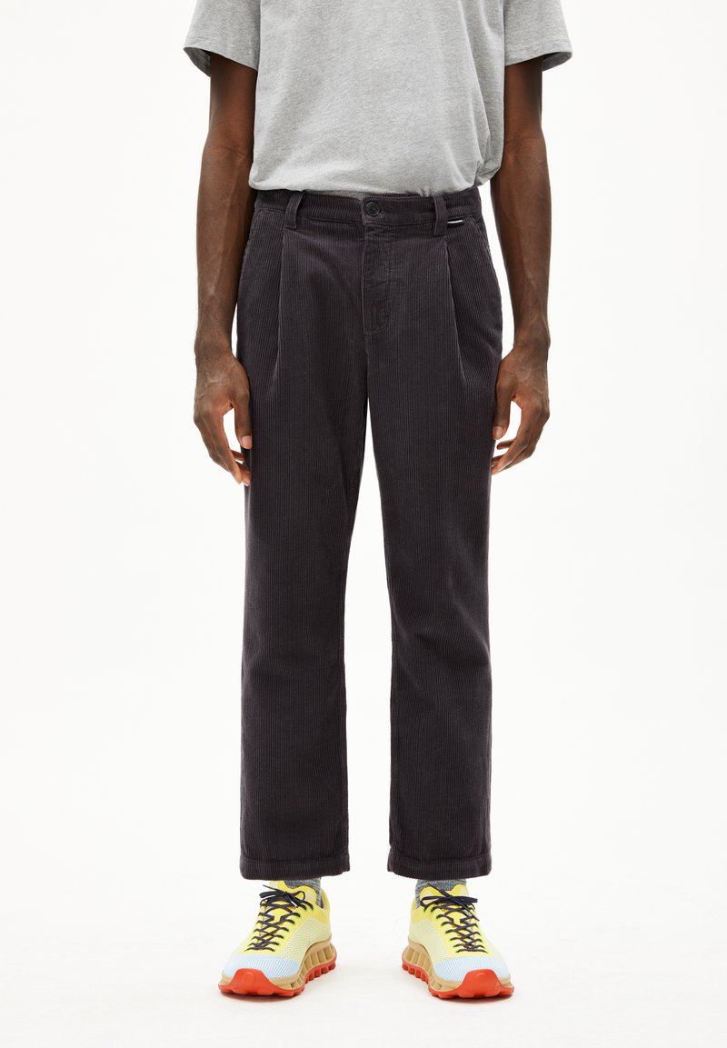 Corduroy broek in donkergrijs met een rechte pijp, plooien aan de voorkant en een knoopsluiting, gecombineerd met levendige gele en oranje sneakers.