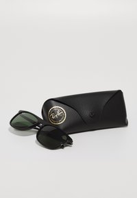 Ochelari de soare Ray-Ban negri cu lentile verzi, cu o formă rectangulară. Însoțiți de un toc texturat negru cu un logo auriu.