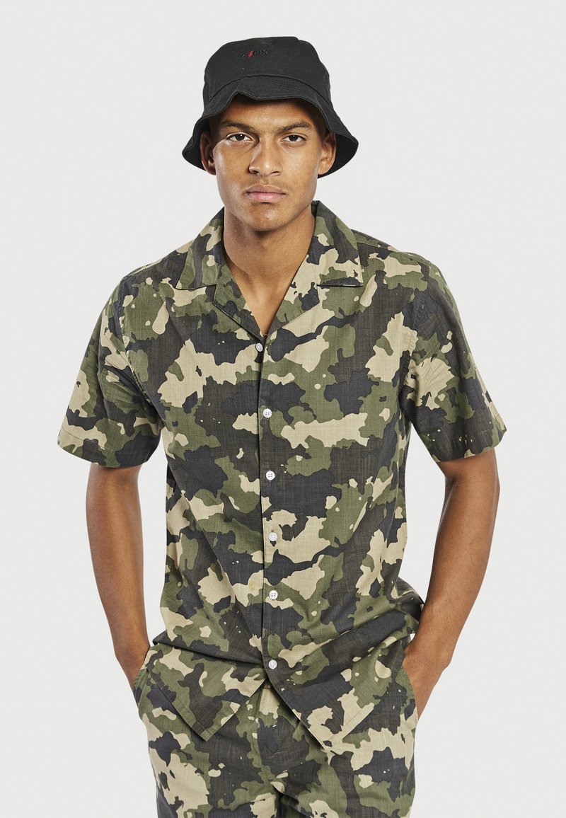 Camisetta a maniche corte con motivo camouflage, caratterizzata da tonalità di verde e nero, con chiusura a bottoni e colletto. Tessuto strutturato, vestibilità comoda.