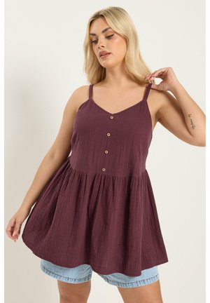 Vrouw die de band van een mouwloze pruimkleurige top met knoopsluiting en uitlopende zoom verstelt, met lichte denimshorts, tegen een effen achtergrond.