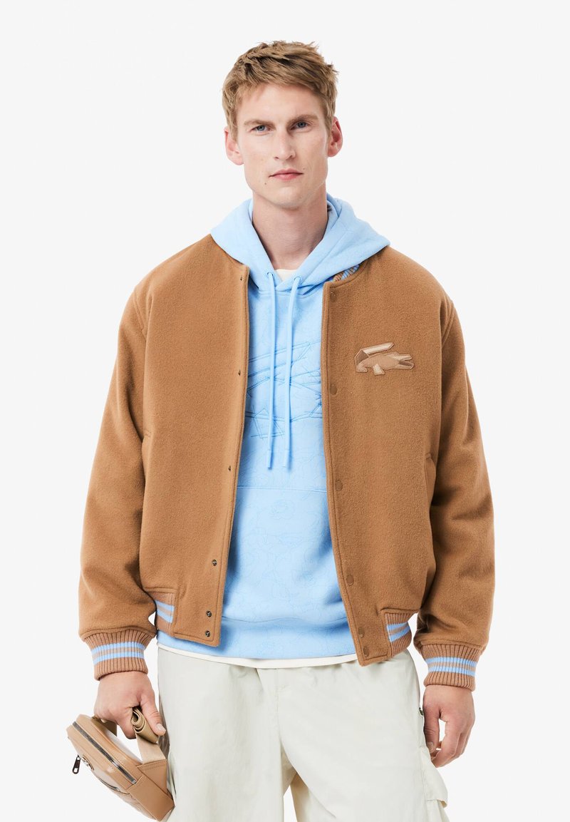 Giacca bomber in fleece marrone con polsini a costine, indossata sopra una felpa con cappuccio azzurro chiaro che presenta un motivo sottile e una tasca a marsupio.