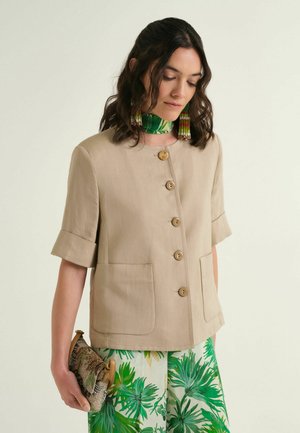 Donna che indossa una giacca beige a maniche corte con bottoni, pantaloni verdi con stampa di foglie, sciarpa verde al collo, lunghi orecchini di perline e tiene una pochette con motivo.