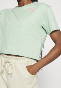 Zelené krátké tričko z měkké látky s kulatým výstřihem. Na levé straně hrudi je růžové logo. Kombinováno s světle krémovými kalhotami, jedna ruka v kapse.