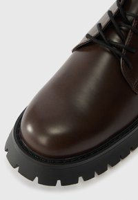 Brauner Lederstiefel mit glatter Oberfläche, ausgestattet mit schwarzen Schnürsenkeln und einer dicken, gerillten schwarzen Sohle mit ausgeprägtem Profil.