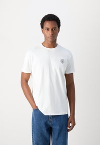 Camiseta de algodón blanca con mangas cortas y cuello redondo. Presenta un pequeño logo bordado en el lado izquierdo del pecho. Combinada con vaqueros de denim azul.