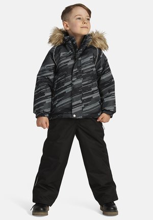 Giacca puffer a motivi neri e grigi con cappuccio foderato in pelliccia, abbinata a pantaloni da neve neri e robuste scarpe nere.