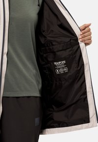Texapore jack met een binnenzak, zwarte nylon voering en een lichte buitenkant, gecombineerd met een groene T-shirt en donkere broeken.