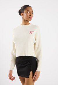 Pulover crem cu nervuri și mâneci lungi, având un design cu fundă roz pe piept. Asortat cu o fustă mini neagră, cu o fanta laterală.