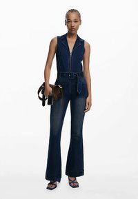 Mono de denim con cuerpo ajustado, cuello y cierre de cremallera, con piernas acampanadas y cintura con cinturón. Color azul oscuro con detalles de costuras.