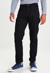 Hombre vestido con pantalones negros con puños, zapatillas altas azul marino y una camisa de rayas blancas y azul marino de manga larga, de pie contra un fondo sencillo.