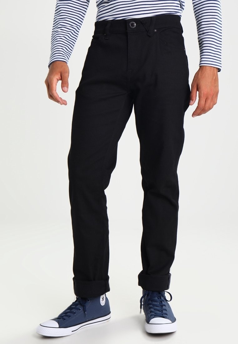 Hombre vestido con pantalones negros con puños, zapatillas altas azul marino y una camisa de rayas blancas y azul marino de manga larga, de pie contra un fondo sencillo.