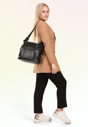 Jonge vrouw in een beige blazer, zwarte broek en witte sneakers, met een grote zwarte leren schoudertas, staande voor een effen achtergrond.