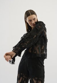 Top din dantelă neagră cu modele florale transparent, mâneci volan și un design scurt. Stilizat cu pantaloni asortați, subliniind textura transparentă.
